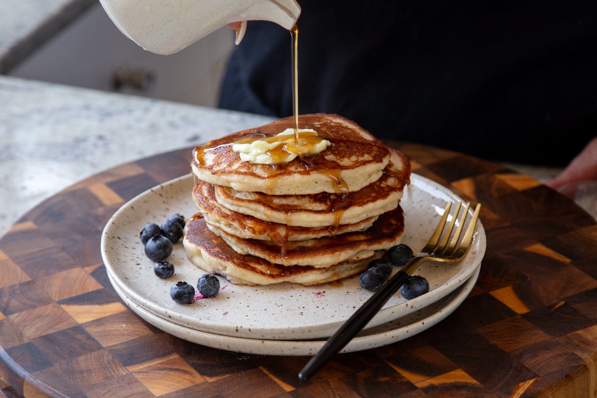 pouring maple syrup onto a stack of gluten-free lemon blueberry pancakes